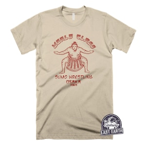 May include: A beige t-shirt featuring a red graphic of a sumo wrestler. The graphic reads "World Class Sumo Wrestling Osaka 大坂府".