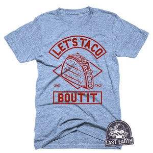 Puede incluir: Una camiseta azul claro con un dise&ntilde;o gr&aacute;fico rojo que dice "Let's Taco Bout It" con una ilustraci&oacute;n roja de un taco en el centro.