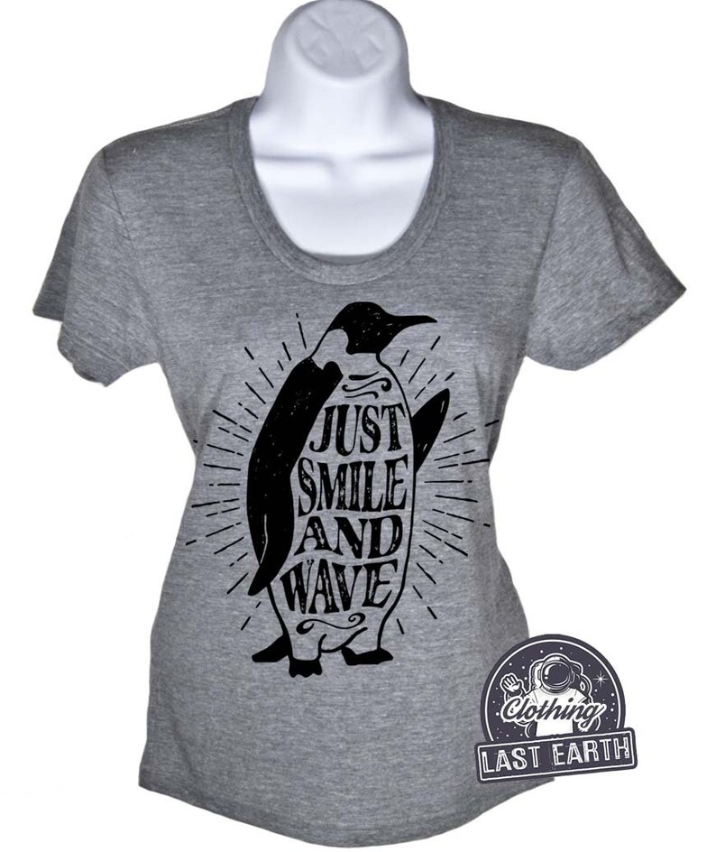 Pu&ograve; includere: Maglietta grigia con un grafico di pinguino nero. Il pinguino tiene una tavola da surf con il testo "Just Smile and Wave" su di essa.