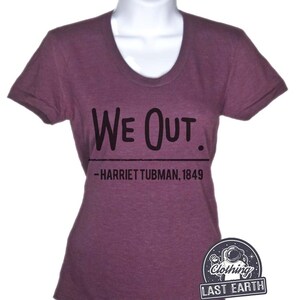Puede incluir: Una camiseta morada con el texto "We Out." y la cita "--Harriet Tubman, 1849" impresa en ella.