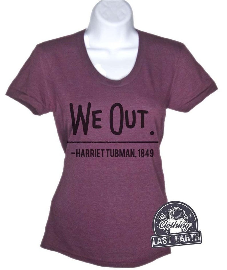 Puede incluir: Una camiseta morada con el texto "We Out." y la cita "--Harriet Tubman, 1849" impresa en ella.