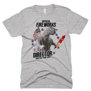 Puede incluir: Una camiseta gris con un gr&aacute;fico de un monstruo parecido a Godzilla que lleva gafas de sol y sostiene un fuego artificial. El gr&aacute;fico est&aacute; rodeado de estrellas rojas, blancas y azules. El texto "OFFICIAL FIREWORKS DIRECTOR RUN FOR YOUR LIFE!" est&aacute; impreso debajo del gr&aacute;fico.