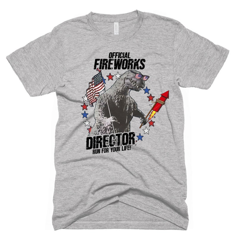 Puede incluir: Una camiseta gris con un gr&aacute;fico de un monstruo parecido a Godzilla que lleva gafas de sol y sostiene un fuego artificial. El gr&aacute;fico est&aacute; rodeado de estrellas rojas, blancas y azules. El texto "OFFICIAL FIREWORKS DIRECTOR RUN FOR YOUR LIFE!" est&aacute; impreso debajo del gr&aacute;fico.