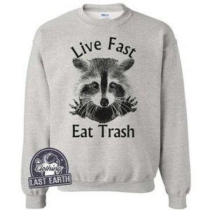 Puede incluir: Una sudadera gris con el texto "Live Fast Eat Trash" impreso en la parte delantera. Una ilustraci&oacute;n en blanco y negro de un mapache asoma por detr&aacute;s del texto.