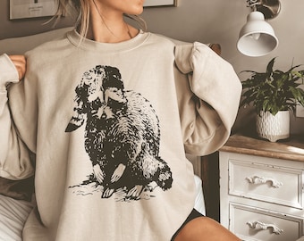 Puede incluir: Una sudadera beige con una ilustración en blanco y negro de un mapache en la parte delantera.