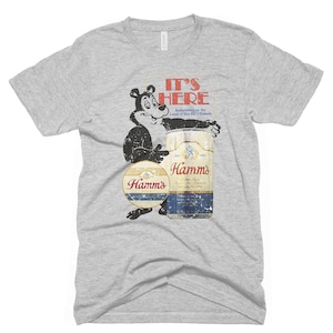 May include: A grey t-shirt with a vintage Hamm's Beer advertisement featuring a cartoon bear holding a can of beer. The text on the advertisement reads "It's Here" and "Refreshing as the Land of Sky Blue Waters".