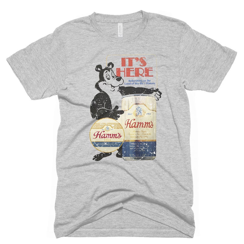 May include: A grey t-shirt with a vintage Hamm's Beer advertisement featuring a cartoon bear holding a can of beer. The text on the advertisement reads "It's Here" and "Refreshing as the Land of Sky Blue Waters".