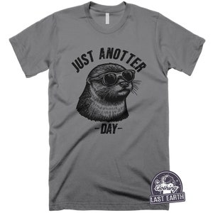 May include: Gray t-shirt with a black and white illustration of an otter wearing sunglasses. The text "Just Anotter Day" is printed on the shirt.
