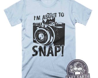 May include: A light blue t-shirt with a black and white graphic of a vintage camera and the text "I'm About To Snap!" printed on the front. The shirt is a unisex style.