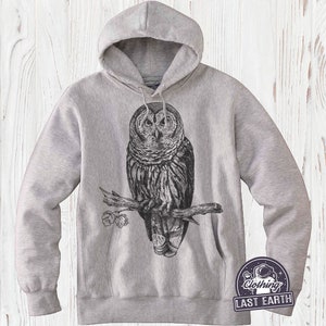 May include: A gray hooded sweatshirt with a black and white illustration of an owl perched on a branch. The owl has large, round eyes and detailed feathers. The sweatshirt has a drawstring hood and a kangaroo pocket.