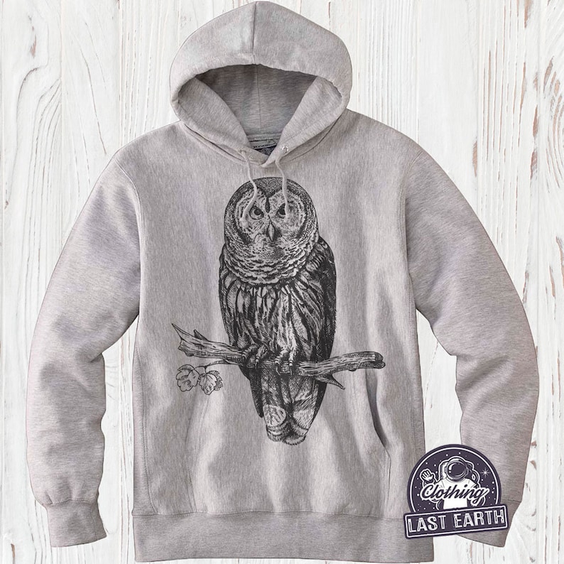 May include: A gray hooded sweatshirt with a black and white illustration of an owl perched on a branch. The owl has large, round eyes and detailed feathers. The sweatshirt has a drawstring hood and a kangaroo pocket.