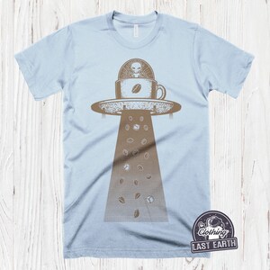May include: A light blue t-shirt with a graphic of a UFO hovering over a coffee cup with an alien inside. Coffee beans are falling from the UFO. The text "Clothing Last Earth" is in a circle with a star and a rocket ship.
