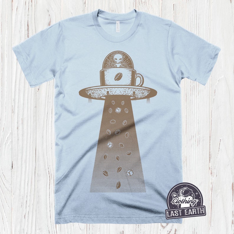 May include: A light blue t-shirt with a graphic of a UFO hovering over a coffee cup with an alien inside. Coffee beans are falling from the UFO. The text "Clothing Last Earth" is in a circle with a star and a rocket ship.