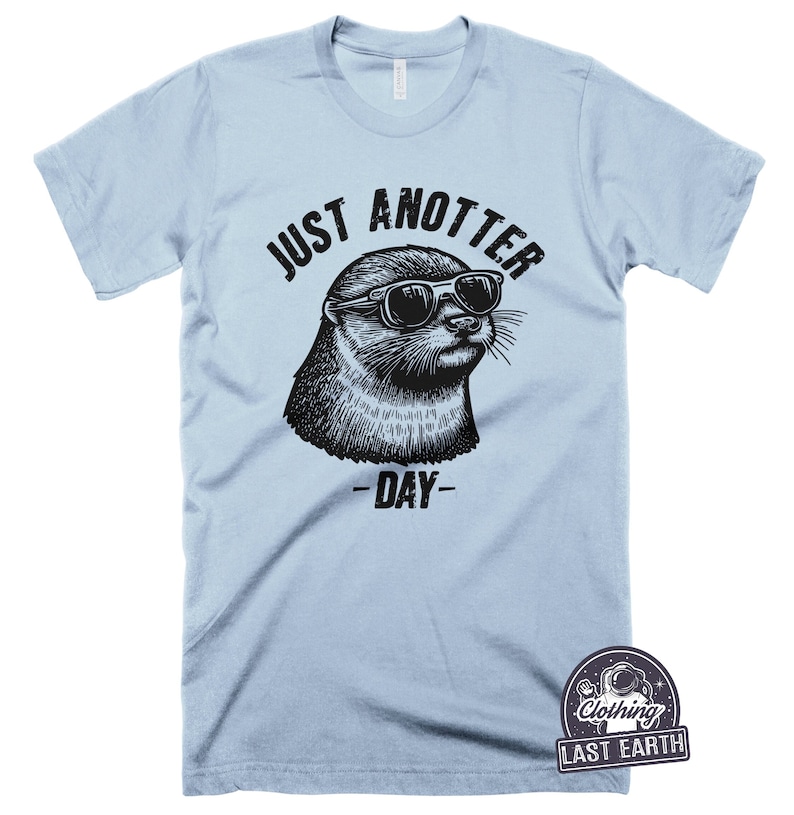 May include: Light blue t-shirt with a black and white illustration of an otter wearing sunglasses. The text "JUST ANOTTER DAY" is printed on the shirt.