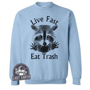 Puede incluir: Sudadera azul claro con una ilustraci&oacute;n en blanco y negro de un mapache asom&aacute;ndose por un agujero con el texto "Live Fast Eat Trash".