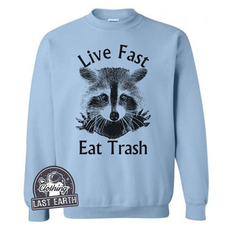 Puede incluir: Sudadera azul claro con una ilustraci&oacute;n en blanco y negro de un mapache asom&aacute;ndose por un agujero con el texto "Live Fast Eat Trash".
