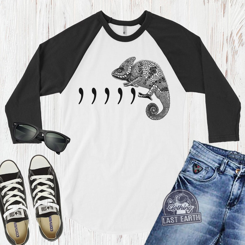 Peut inclure: Un t-shirt de baseball blanc et noir avec un graphique de cam&eacute;l&eacute;on noir et le texte ",,,,," imprim&eacute; sur le devant. Le t-shirt a des manches noires et un corps blanc.