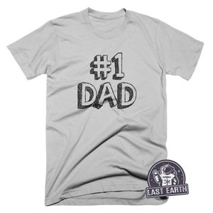 May include: A light gray t-shirt with the text "#1 DAD" in black. The text is in a hand-drawn style. The shirt is a standard crew neck t-shirt.