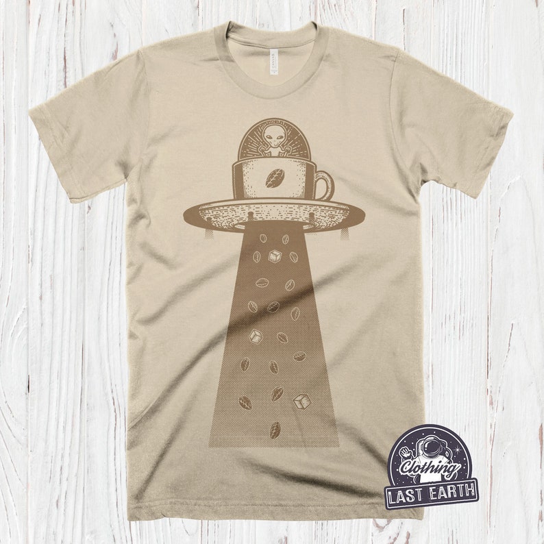 May include: A beige t-shirt with a graphic of a UFO shaped like a coffee cup with an alien inside. Coffee beans are falling from the UFO.