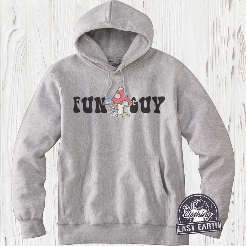 K&ouml;nnte beinhalten: Ein grauer Kapuzenpullover mit dem Text "FUN GUY" in schwarzen Buchstaben und einer Cartoon-Pilzgrafik.