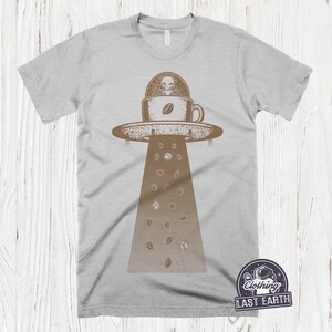 May include: A gray t-shirt with a graphic of a UFO holding a coffee cup with an alien inside. Coffee beans and sugar cubes fall from the UFO. The text "Last Earth" is on the bottom right corner of the shirt.
