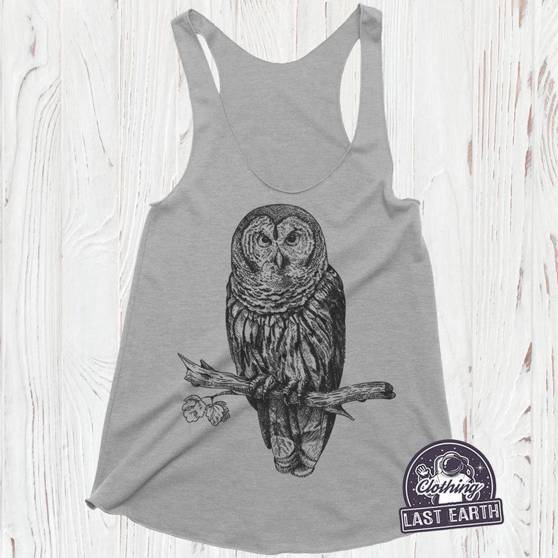May include: Gray tank top with a black and white illustration of an owl perched on a branch. The owl is looking directly at the viewer. The text "Clothing Last Earth" is printed on the bottom right corner of the tank top.