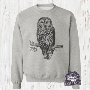 May include: A gray sweatshirt with a black and white illustration of an owl perched on a branch. The owl has a detailed design with feathers and a sharp beak. The sweatshirt has a crew neck and long sleeves.