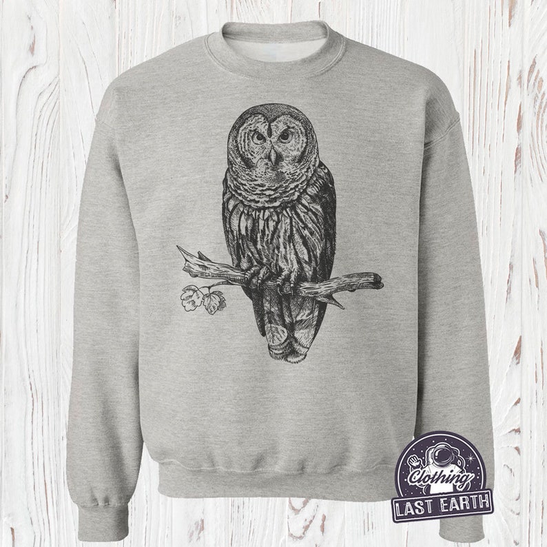 May include: A gray sweatshirt with a black and white illustration of an owl perched on a branch. The owl has a detailed design with feathers and a sharp beak. The sweatshirt has a crew neck and long sleeves.