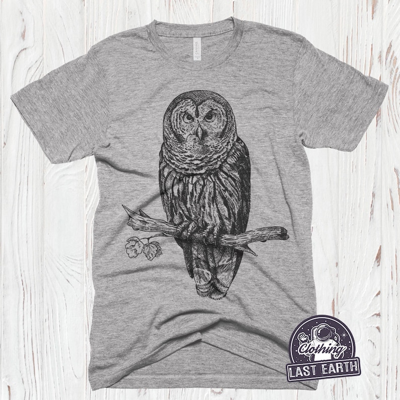 May include: A heather grey t-shirt with a black and white illustration of an owl perched on a branch. The owl has a detailed design with feathers and a serious expression. The t-shirt has a small logo in the bottom right corner that says "Clothing Last Earth".