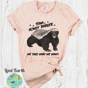 May include: A pink t-shirt with a black and white illustration of a honey badger. The shirt has the text "Team Honey Badger" and "We take what we want!" printed on it.
