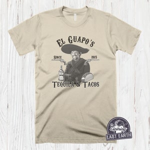 K&ouml;nnte beinhalten: Ein beiges T-Shirt mit einem schwarzen Grafikdruck eines Mannes mit Schnurrbart und Sombrero, der eine Flasche Tequila und einen Taco h&auml;lt. Der Text auf der Grafik lautet "El Guapo's Since 1915 Tequila & Tacos".