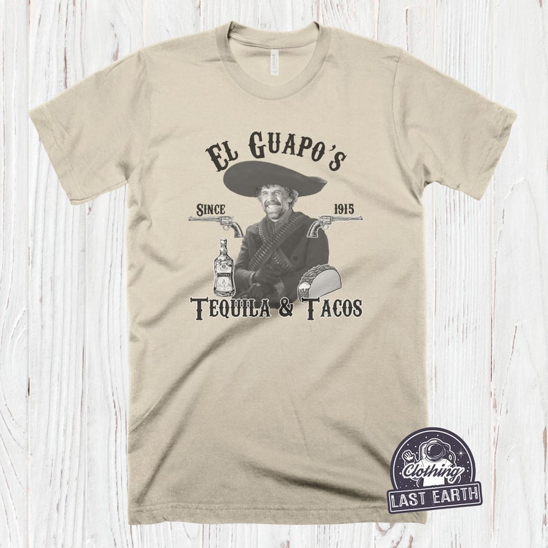 K&ouml;nnte beinhalten: Ein beiges T-Shirt mit einem schwarzen Grafikdruck eines Mannes mit Schnurrbart und Sombrero, der eine Flasche Tequila und einen Taco h&auml;lt. Der Text auf der Grafik lautet "El Guapo's Since 1915 Tequila & Tacos".