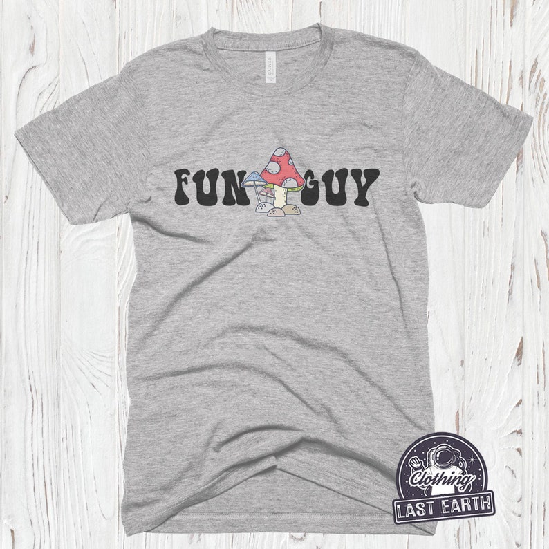 K&ouml;nnte beinhalten: Ein heidefarbenes T-Shirt mit dem Text "FUN GUY" in schwarzen Buchstaben. Ein Cartoon-Bild von drei Pilzen ist zwischen den W&ouml;rtern zentriert.
