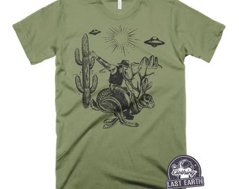 May include: A green t-shirt with a black and white graphic print of a cowboy riding a jackrabbit. The graphic also includes a cactus, a sun, and two flying saucers. The t-shirt has the text "Last Earth Clothing" printed on the bottom right corner.