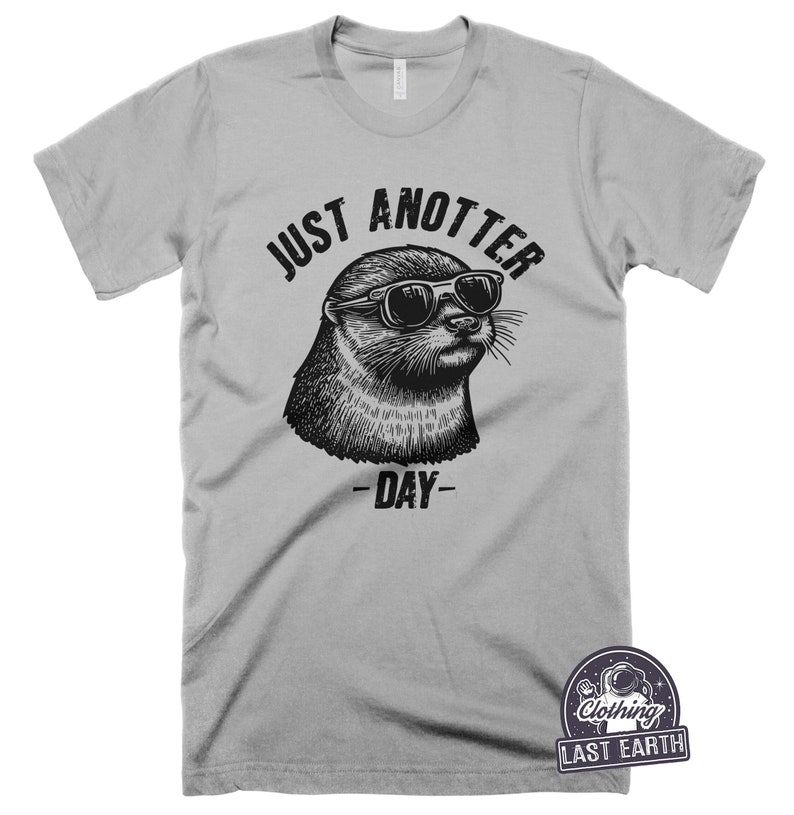 May include: A gray t-shirt with a black and white illustration of an otter wearing sunglasses. The text "Just Anotter Day" is printed on the shirt.