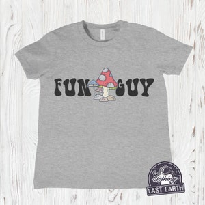 K&ouml;nnte beinhalten: Ein heidefarbenes T-Shirt mit dem Text "FUN GUY" in schwarzer, Retro-Schrift. Ein Cartoon-Bild von drei Pilzen ist mittig zwischen den W&ouml;rtern platziert.