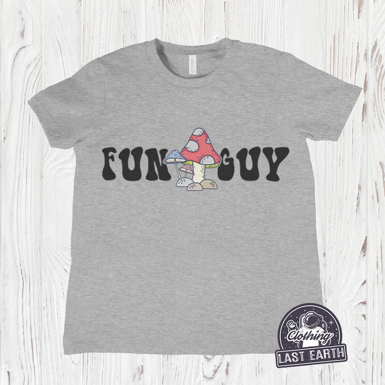 K&ouml;nnte beinhalten: Ein heidefarbenes T-Shirt mit dem Text "FUN GUY" in schwarzer, Retro-Schrift. Ein Cartoon-Bild von drei Pilzen ist mittig zwischen den W&ouml;rtern platziert.