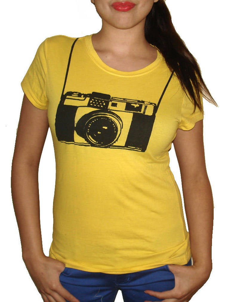 May include: A yellow t-shirt with a black and white graphic of a camera with a strap.