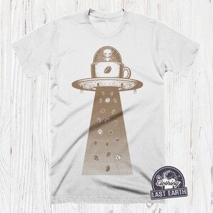 May include: White t-shirt with a brown graphic of a flying saucer with an alien inside a coffee cup. Coffee beans and sugar cubes are falling from the saucer.
