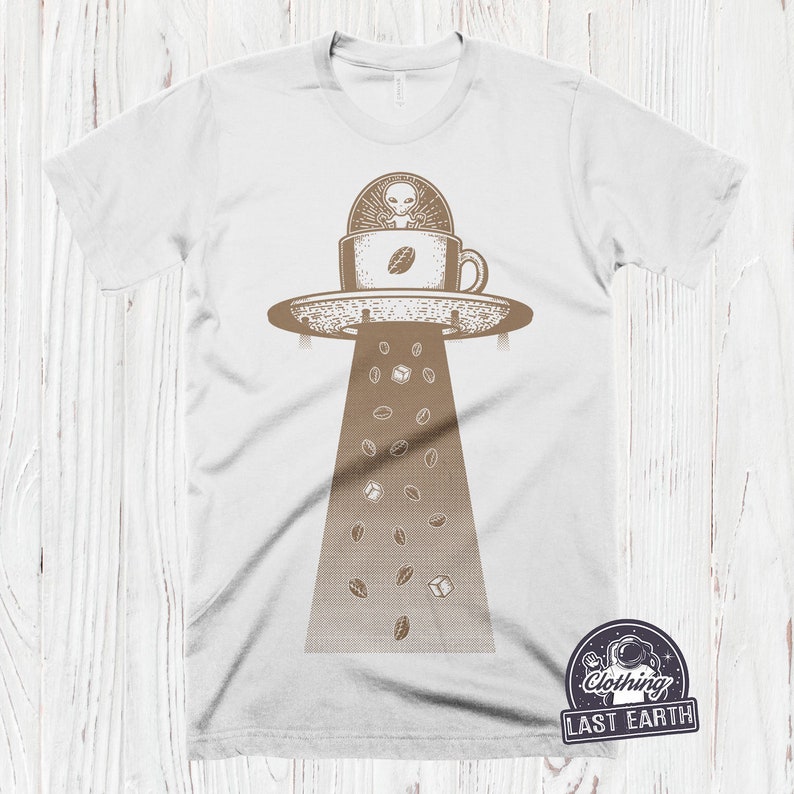 May include: White t-shirt with a brown graphic of a flying saucer with an alien inside a coffee cup. Coffee beans and sugar cubes are falling from the saucer.