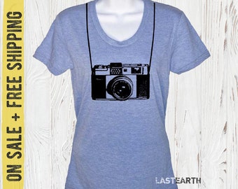 May include: A light blue t-shirt with a black graphic of a vintage camera with a strap. The text "LASTEARTH" is printed on the bottom of the shirt.