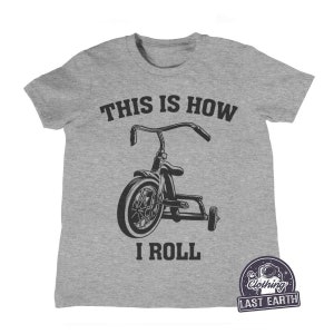Può includere: Una maglietta grigia con il testo "This is how I roll" e un'illustrazione in bianco e nero di un triciclo.