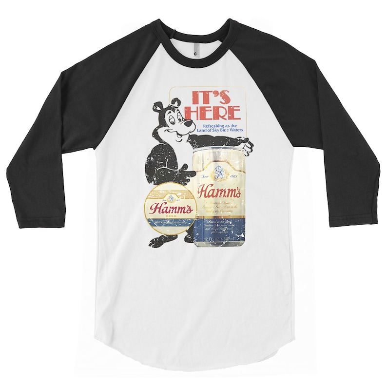 May include: A white and black baseball style t-shirt with a vintage Hamm's Beer advertisement featuring a cartoon bear holding a can of beer. The text on the advertisement reads "It's Here" and "Refreshing as the Land of Sky Blue Waters".