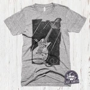 May include: A grey t-shirt featuring a black and white graphic of a skeleton playing an electric guitar. The graphic is in a rectangular frame with a distressed look. The t-shirt has the text "Clothing Last Earth" in a circle with a star and an astronaut graphic.