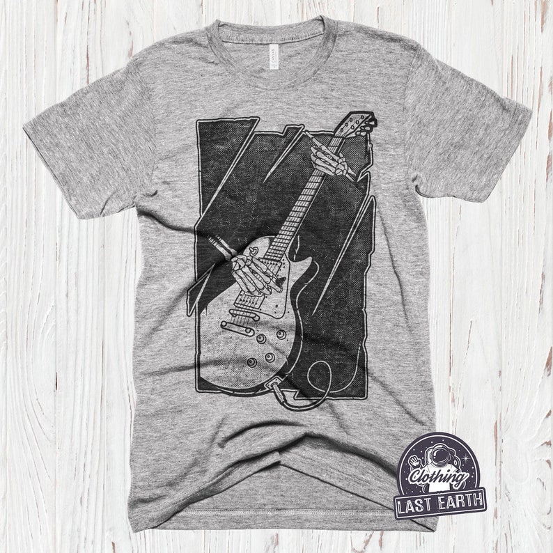 May include: A grey t-shirt featuring a black and white graphic of a skeleton playing an electric guitar. The graphic is in a rectangular frame with a distressed look. The t-shirt has the text "Clothing Last Earth" in a circle with a star and an astronaut graphic.