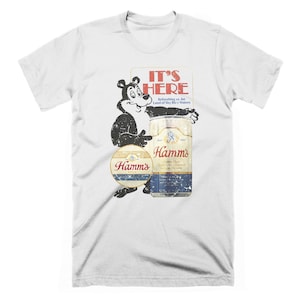 May include: White t-shirt with a vintage-style graphic of a bear holding a can of Hamm's beer. The text "It's Here" is above the bear and can, with the tagline "Refreshing as the Land of Sky Blue Waters".