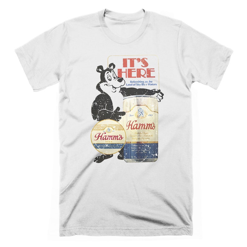 May include: White t-shirt with a vintage-style graphic of a bear holding a can of Hamm's beer. The text "It's Here" is above the bear and can, with the tagline "Refreshing as the Land of Sky Blue Waters".