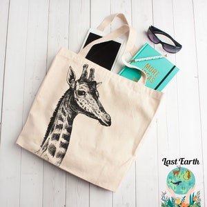 May include: A beige canvas tote bag with a black and white illustration of a giraffe's head. The bag has black handles and is filled with a tablet, a turquoise notebook with the text "More Ideas" on the cover, and sunglasses.