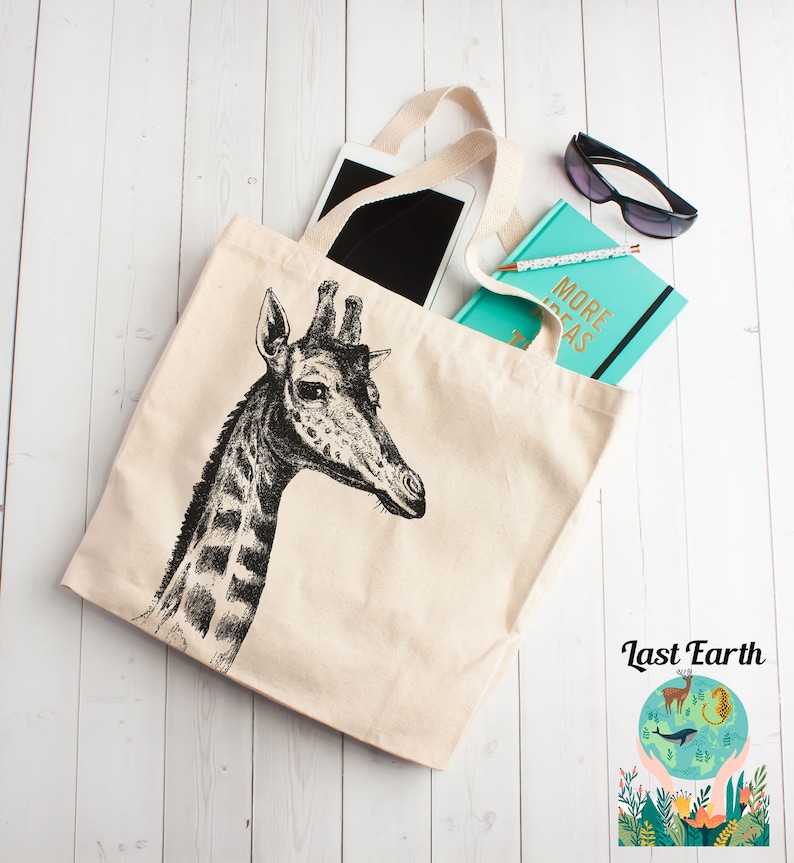 May include: A beige canvas tote bag with a black and white illustration of a giraffe's head. The bag has black handles and is filled with a tablet, a turquoise notebook with the text "More Ideas" on the cover, and sunglasses.