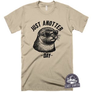 May include: A tan t-shirt with a black graphic of an otter wearing sunglasses and the text "Just Anotter Day".